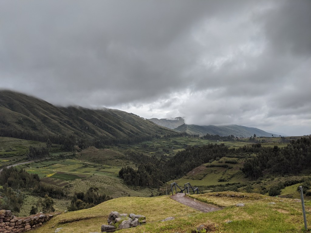 The view from Puka Pukara, including rolling hills and mountains under rain clouds