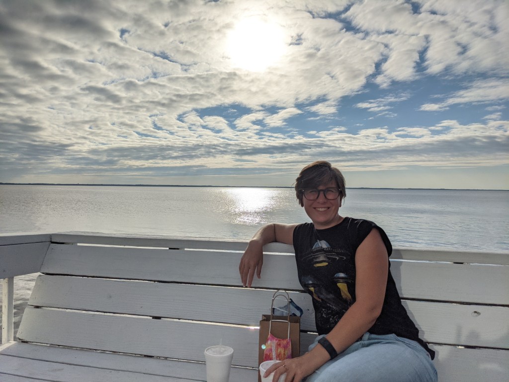 Mel sits on a white bench in front of calm water reflecting white clouds and sunshine.
