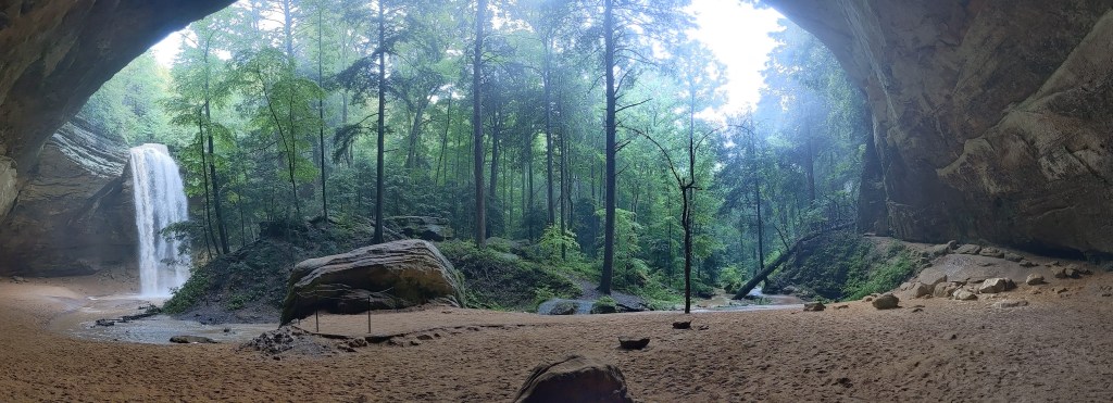 A panoramic shot with a large cave opening framing a waterfall on the left, moss-covered oasis in the center with tall green trees, and sand all around.