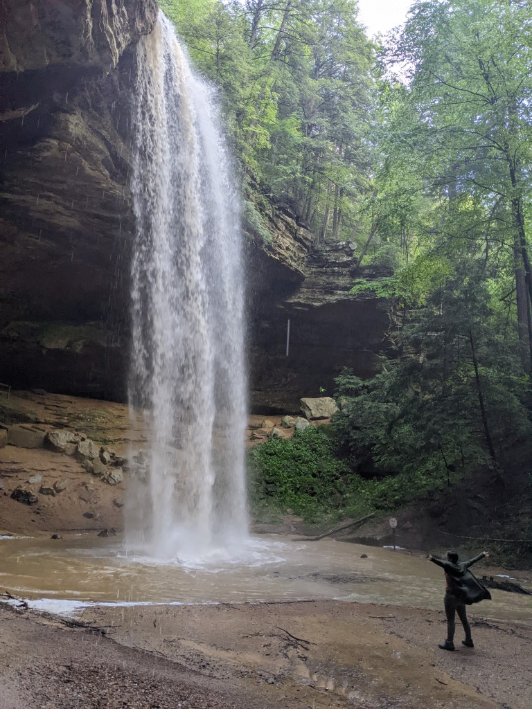 A waterfall pours onto the ground, creating a water basin, while the Scaredy Cat stands below, arms up and wide, with her jacket blowing in the wind created by the fall. 