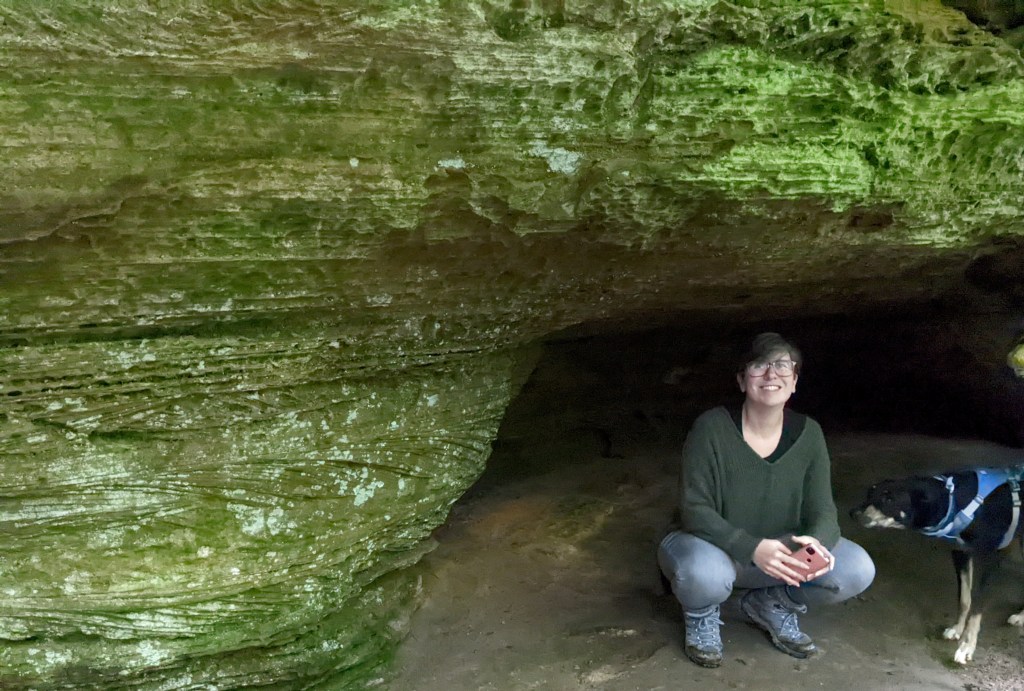 The Scaredy Cat, wearing a green sweater, crouches in a small cave in a lichen-covered rock. A black dog in a blue vest sniffs at her arm.